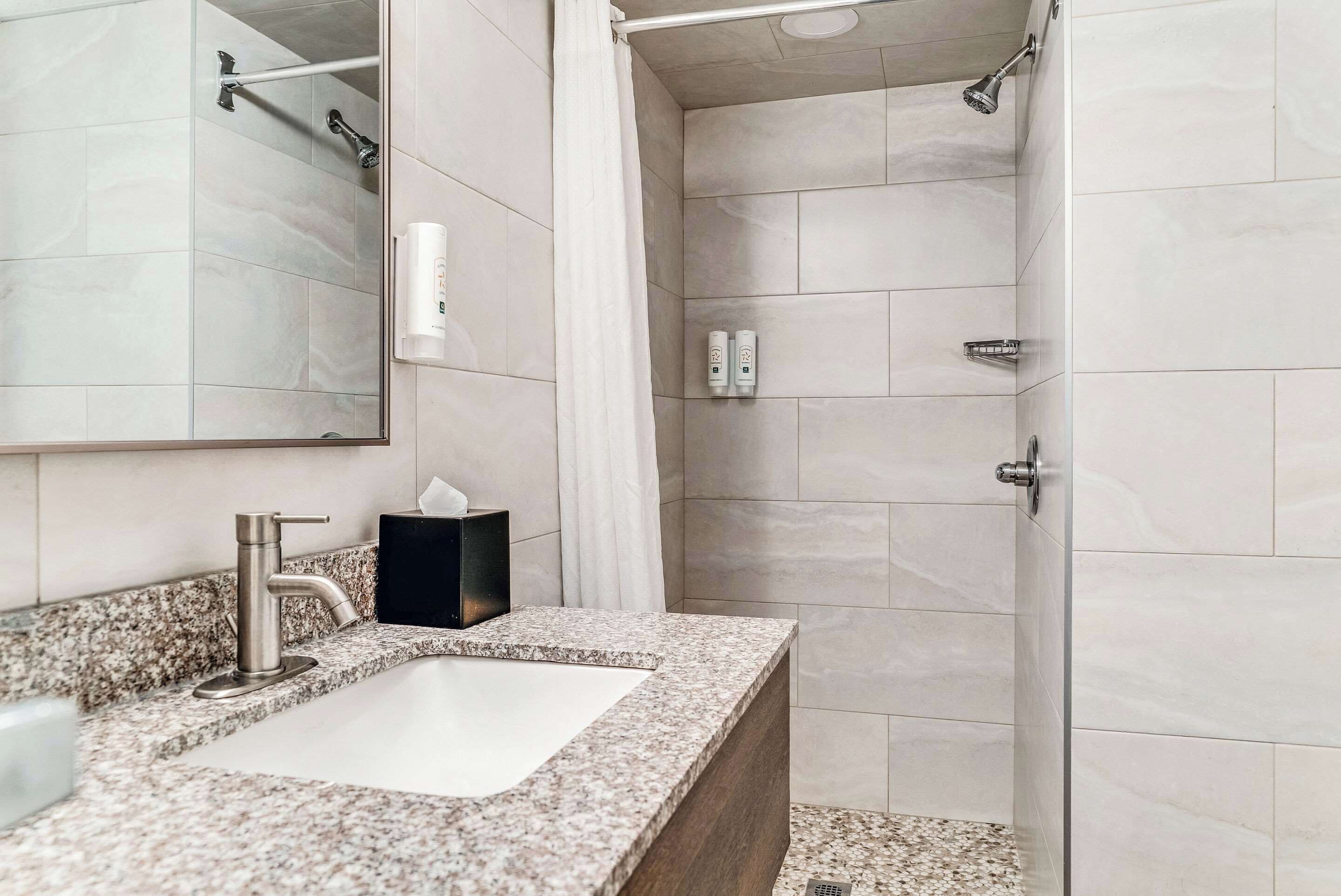 Un baño moderno con encimera de granito, un lavabo blanco, un espejo y una zona de ducha con puerta de cristal.