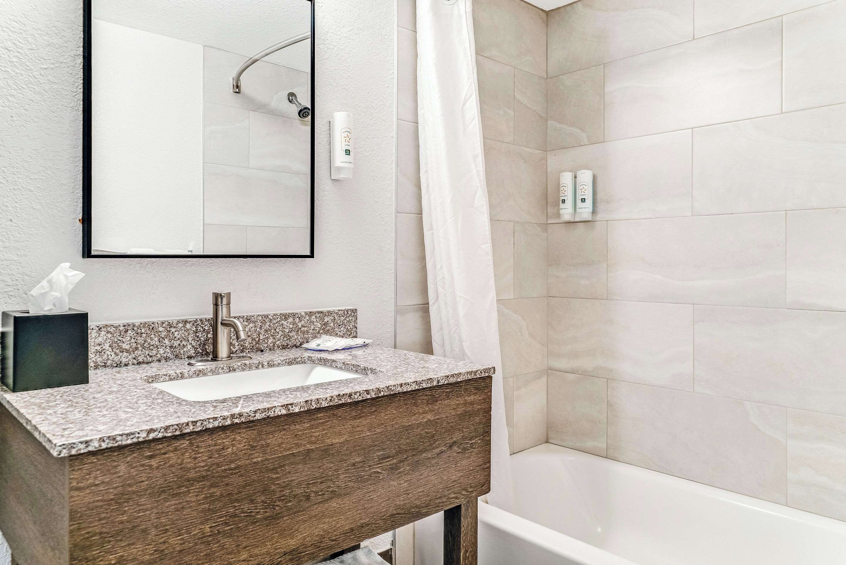 A bathroom with a granite countertop sink, a wooden vanity, a mirror, a shower with a curtain, and a tiled wall.