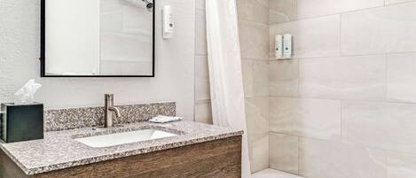 A bathroom with a granite countertop sink, a wooden vanity, a mirror, a shower with a curtain, and a tiled wall.
