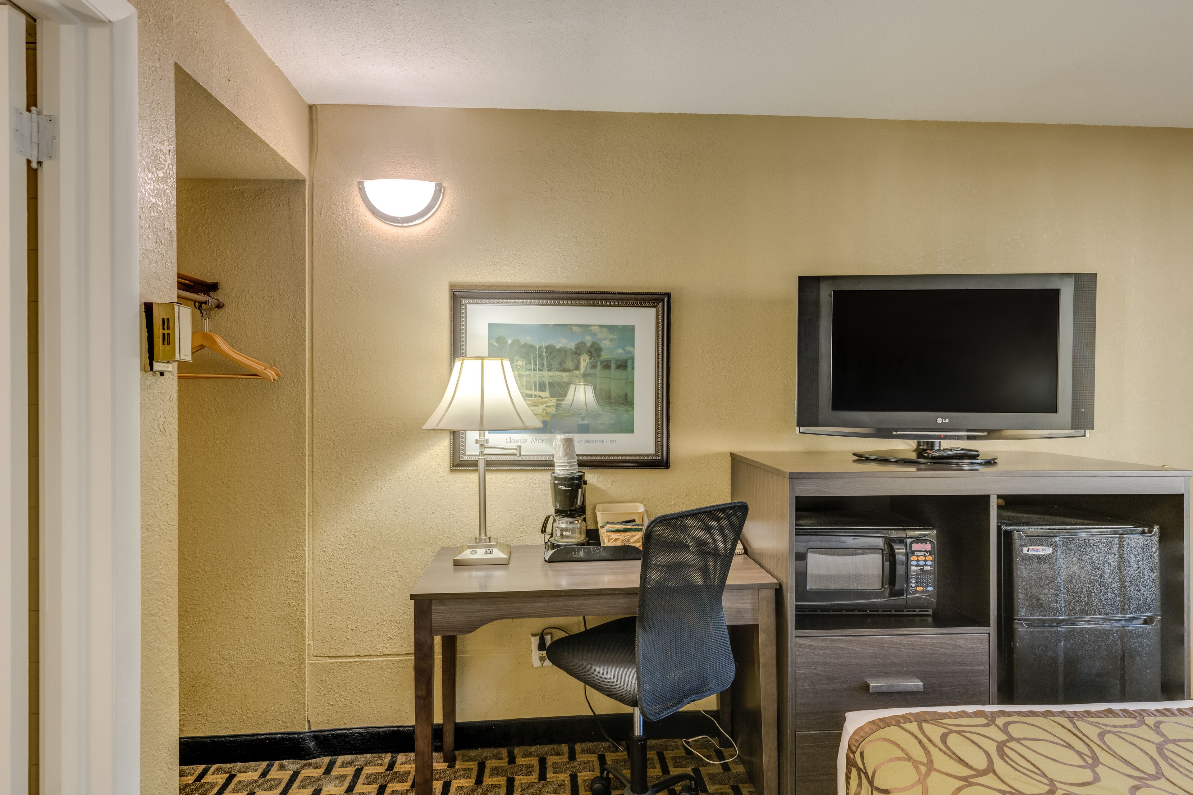 Quarto de hotel com escrivaninha, cadeira, televisão, micro-ondas, frigorífico e uma cama com roupa de cama estampada.