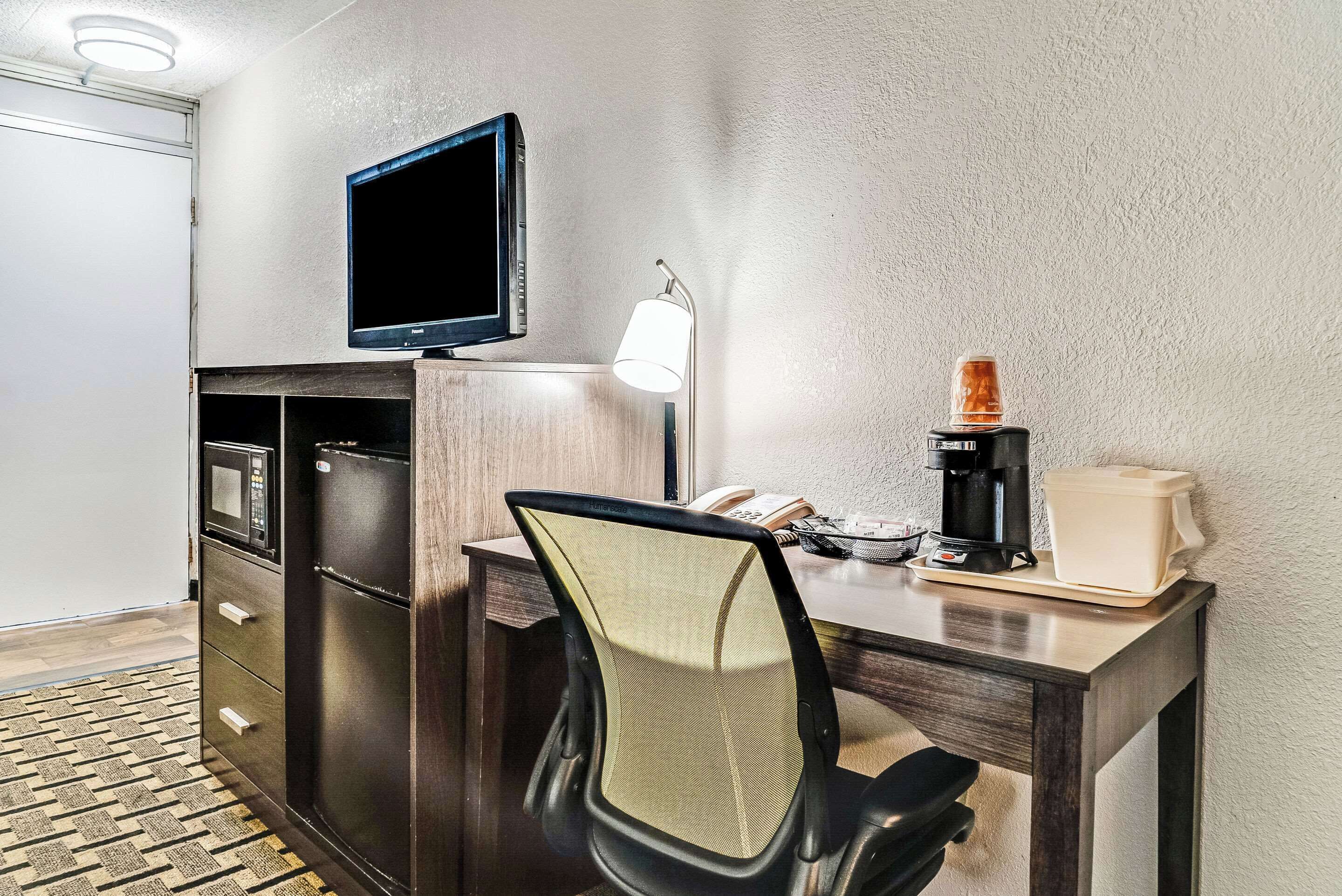 Quarto de hotel compacto com escrivaninha de madeira escura, televisão montada acima, luminária, cafeteira e geladeira.