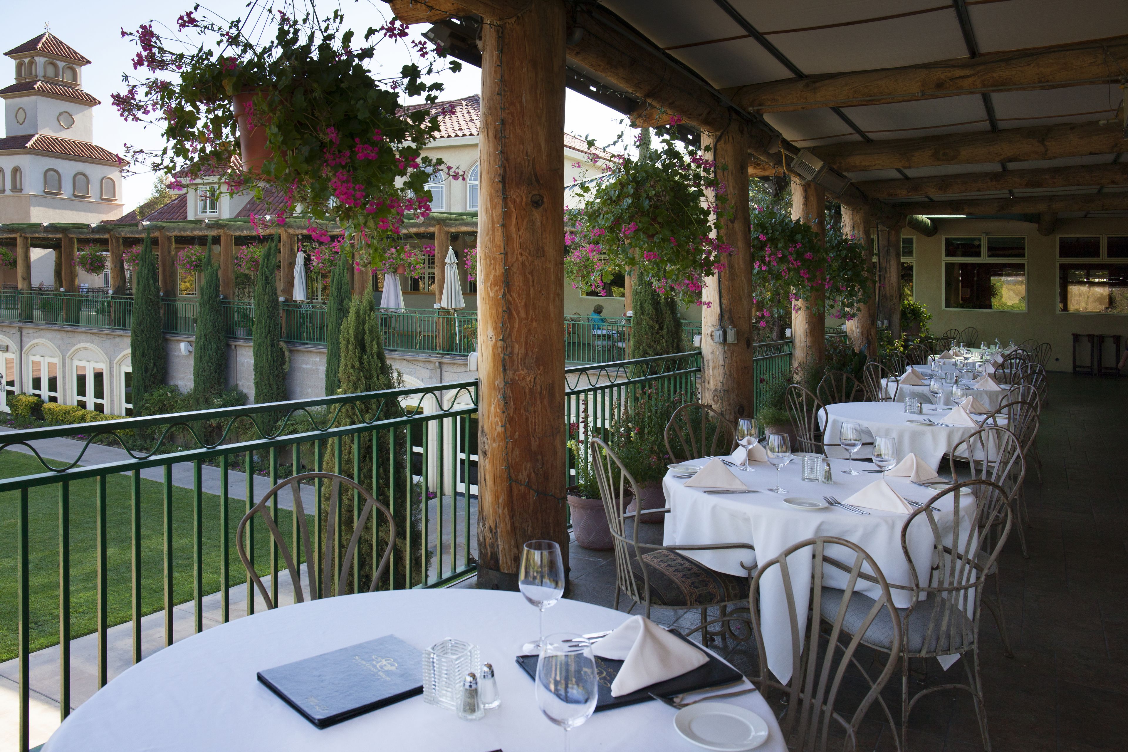 An outdoor dining area with tables set for a meal, surrounded by greenery and a view of a building with a tower.