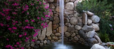Une cascade d’eau se déversant sur un mur de pierre et se jetant dans un étang, le tout entouré d’une végétation luxuriante et de fleurs roses.