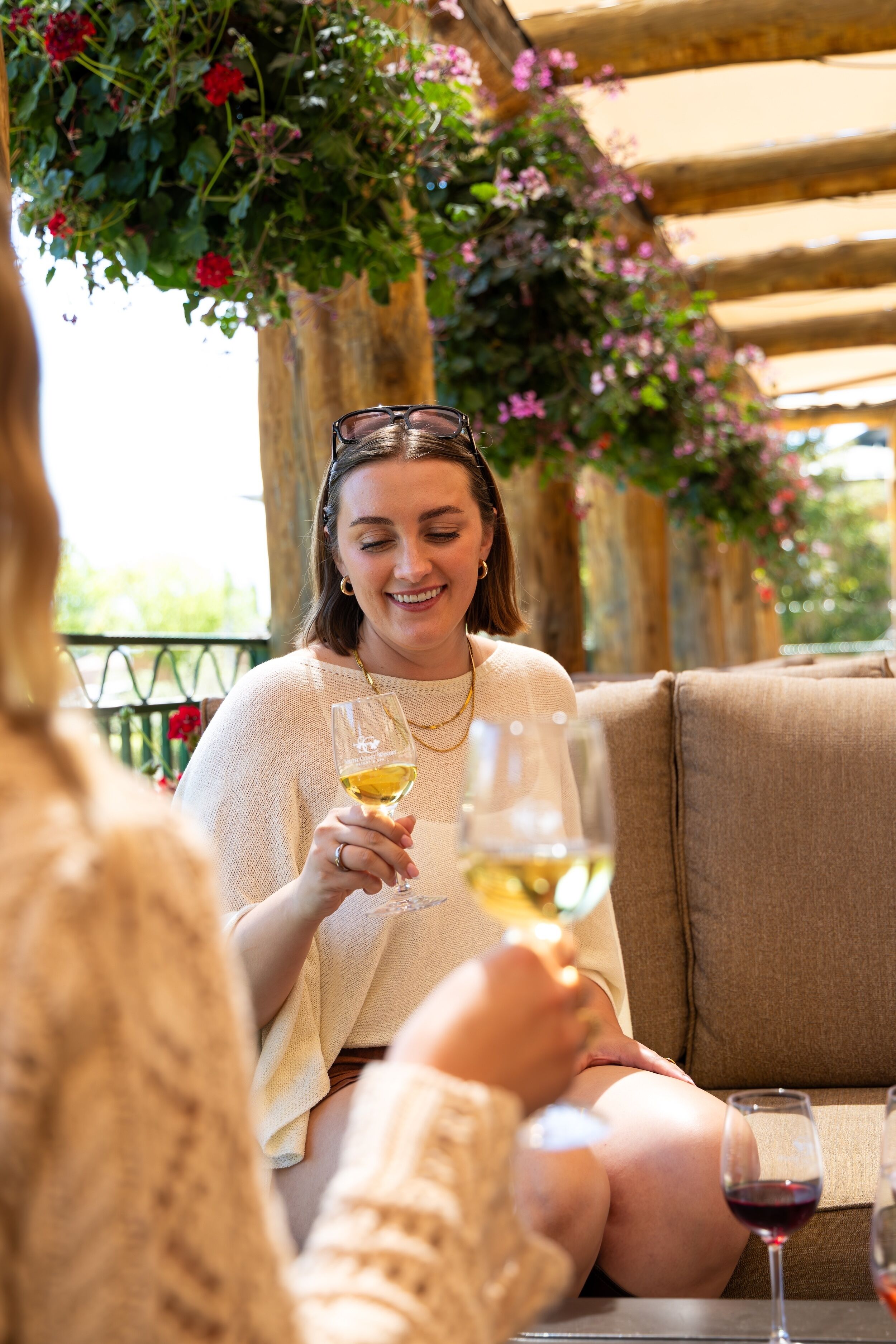 Une personne est assise sur un canapé beige, un verre de vin blanc à la main, entourée de verdure et d’une structure en bois.