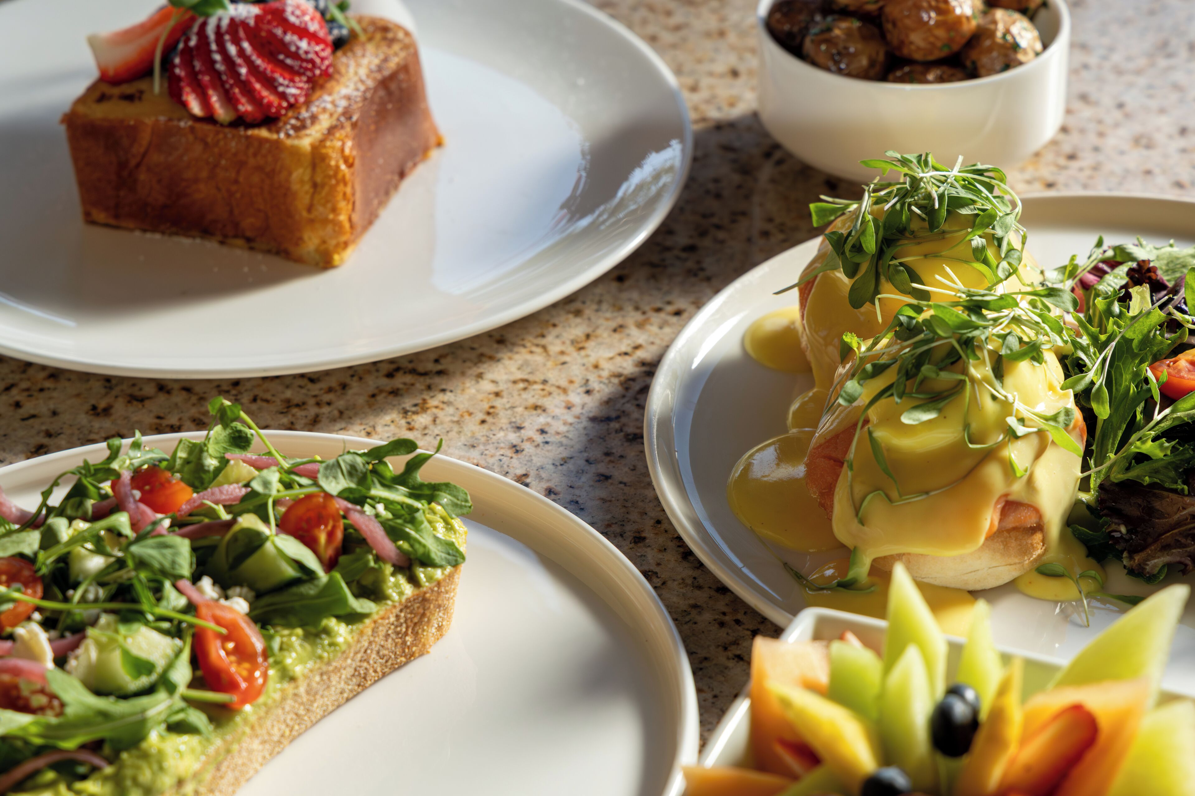 A variety of plated dishes including a slice of cake with strawberries, a salad with greens and tomatoes, and a dish with poached eggs and greens.