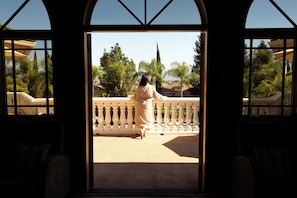 A woman standing on a balcony with a view of palm trees and a building in the background.