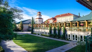 A courtyard with a building featuring a tower, a covered walkway with hanging flower baskets, and a well-maintained lawn. - South Coast Winery Resort and Spa (Temecula)