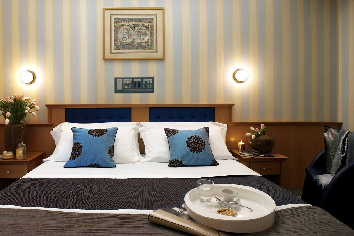 A hotel room with a bed, a nightstand, a tray with a glass of water, a vase with flowers, and a framed picture on the wall.