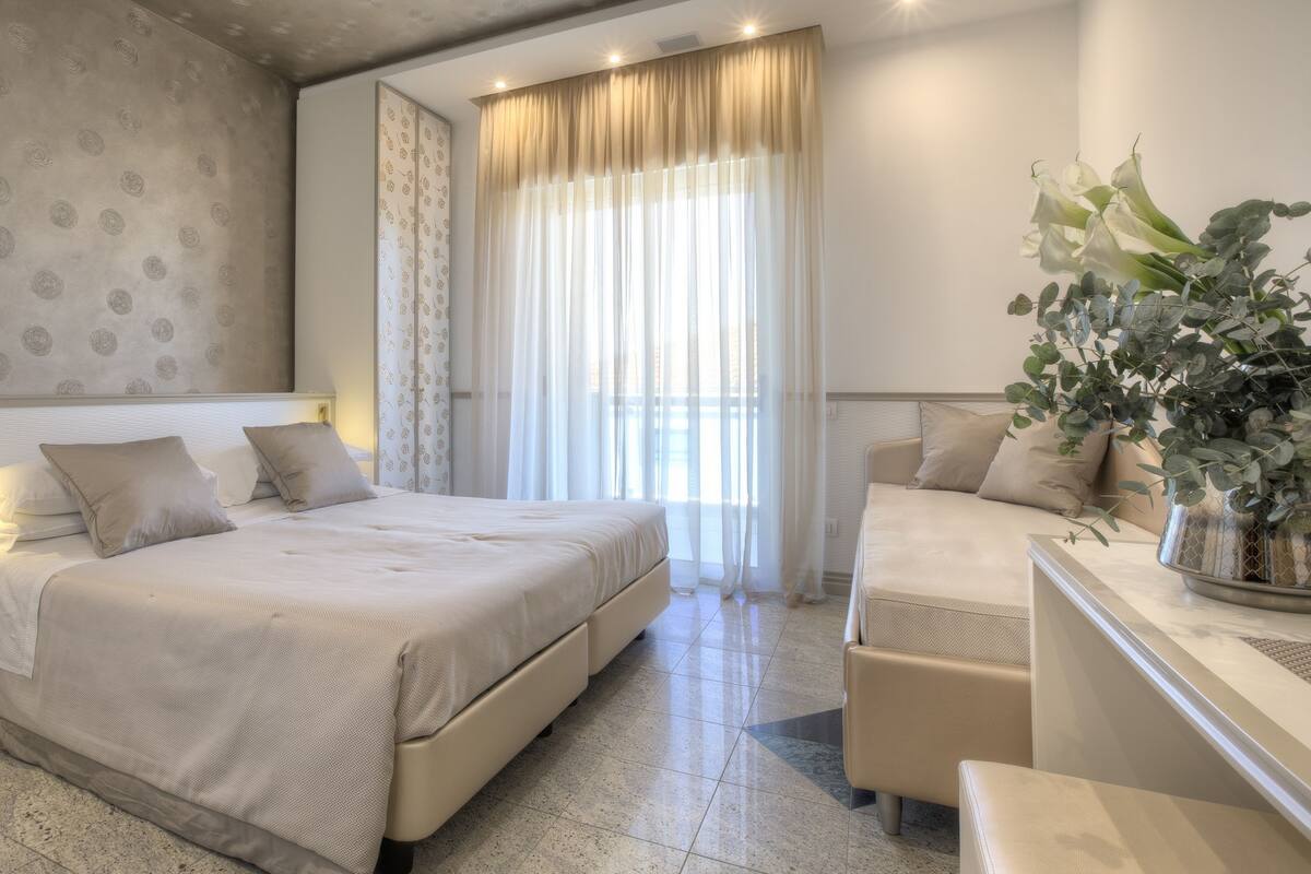 A hotel room with a bed, a sofa, a potted plant, and a window with curtains.