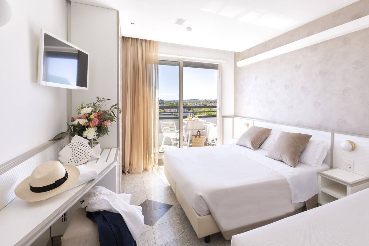 A hotel room with two beds, a desk with a chair, a TV, and a balcony with a table and chairs.