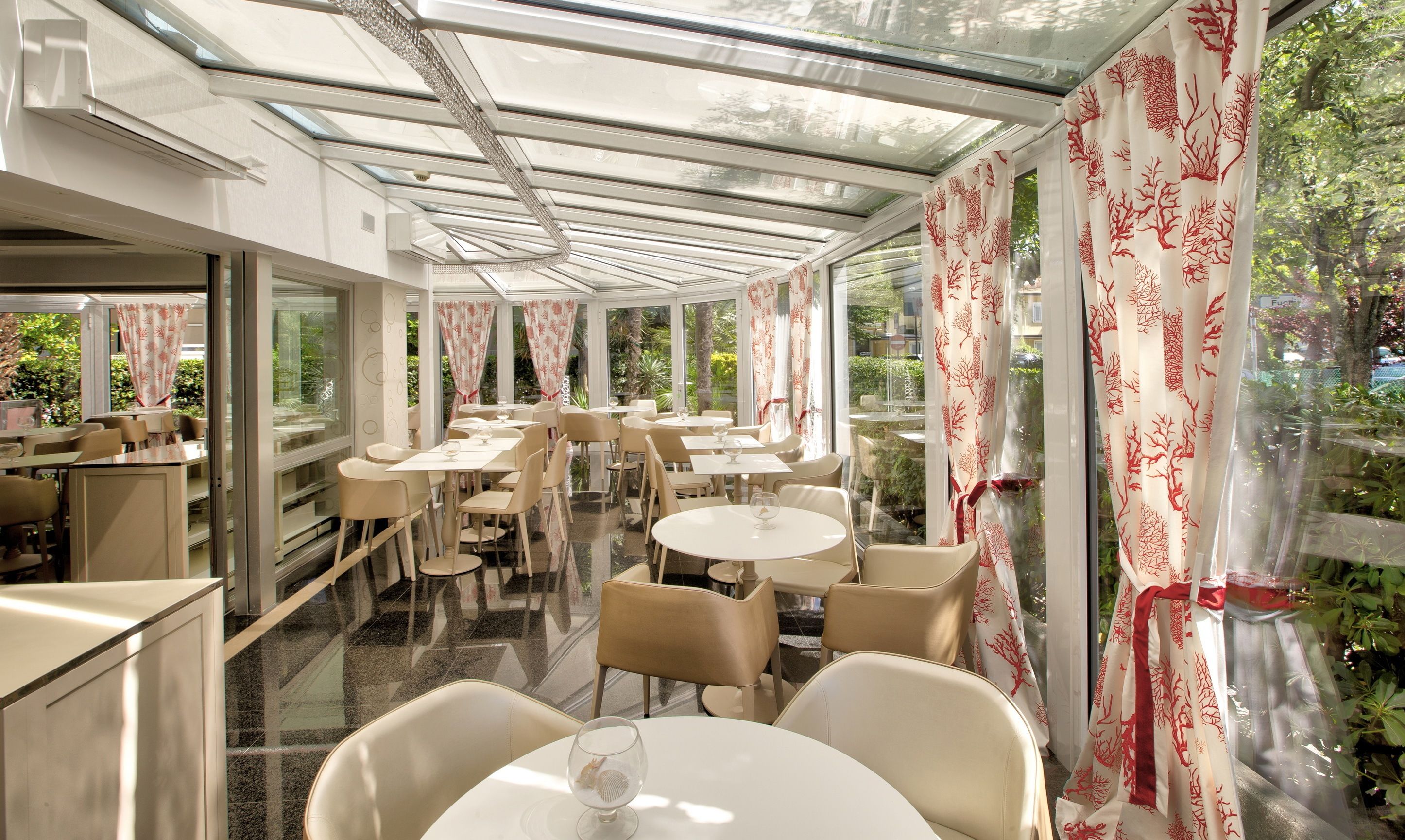Ein Restaurant mit Glasdecke, weißen Tischen und Stühlen sowie roten, gemusterten Vorhängen.