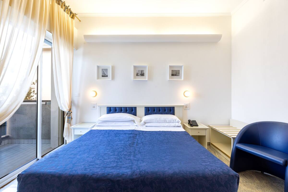 A hotel room with a large bed, a desk, a chair, and a balcony with curtains.