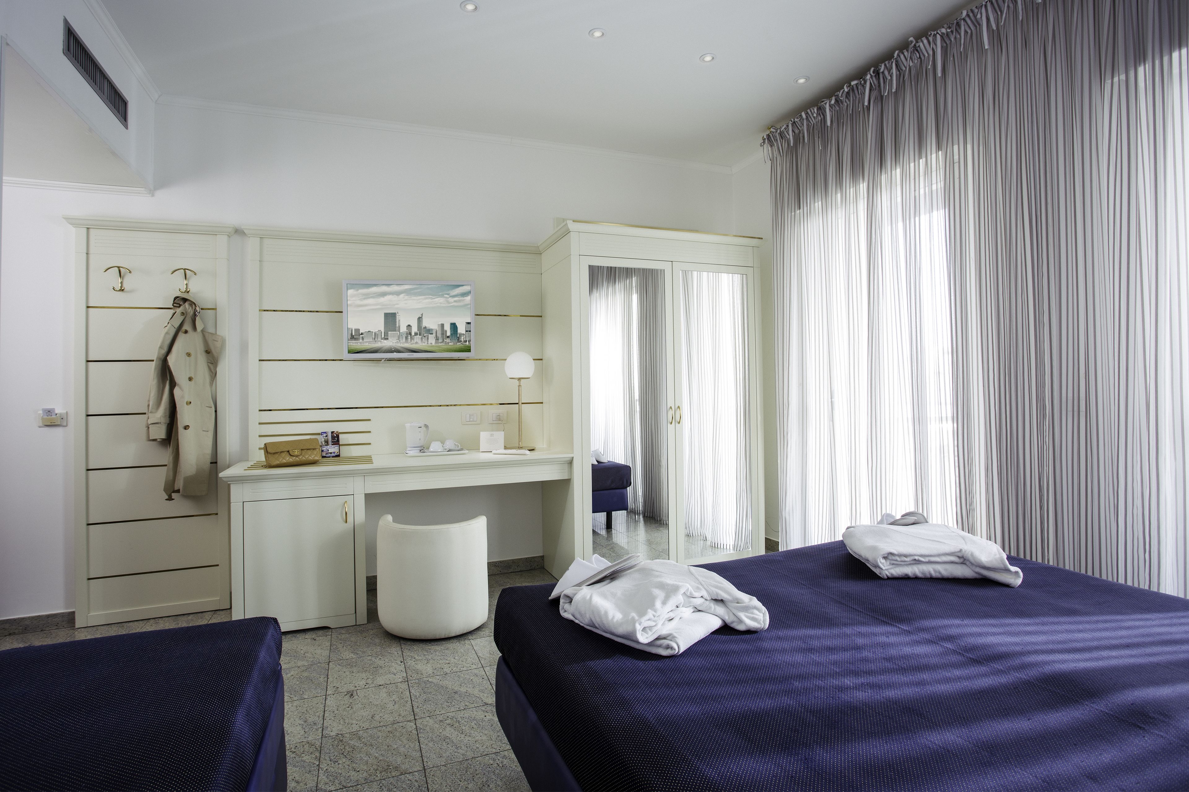 Habitación de hotel con dos camas, un escritorio, una silla, un armario y un ventanal con cortinas.