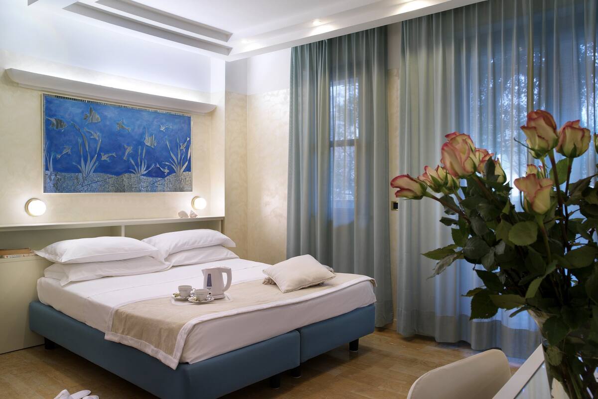 A modern hotel room with a large bed, a bedside table with a lamp, a painting on the wall, and a window with curtains.