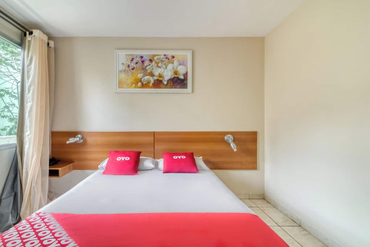 A hotel room with a bed, a headboard, a nightstand, a window with curtains, and a wall-mounted painting.