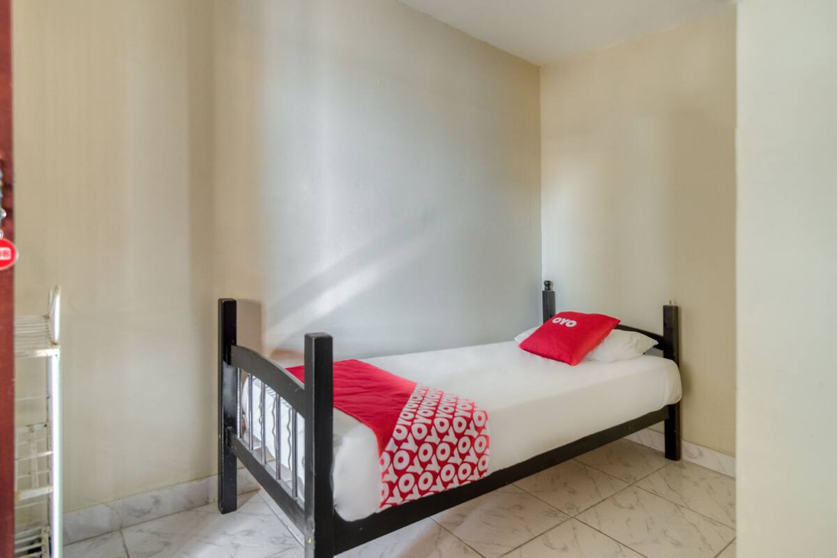 A single bed with a patterned bedspread and red pillows in a room with tiled flooring and plain walls.