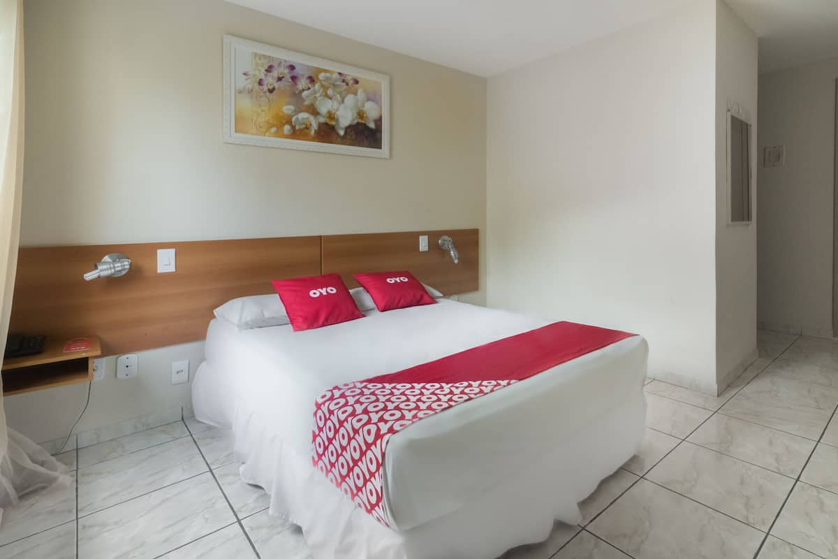 A hotel room with a bed, a headboard, a bedside table, a nightstand, a wall-mounted artwork, and a tiled floor.