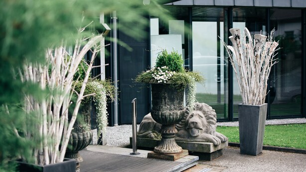 A modern building with large windows, a stone lion sculpture, and various potted plants. - Fritz am Brunnen (Schwelm)