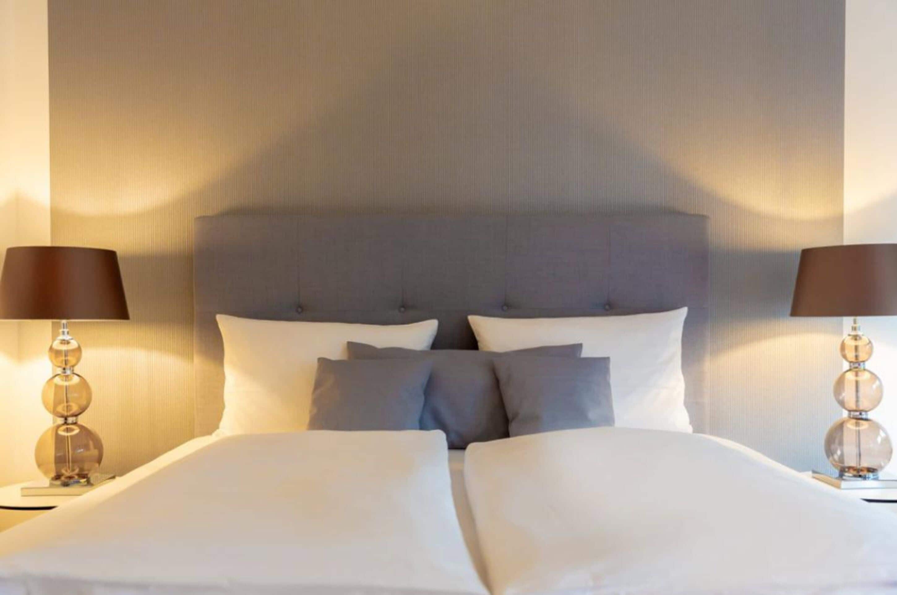 Una cama bien hecha con un cabecero gris, dos lámparas de noche y una pared con textura.