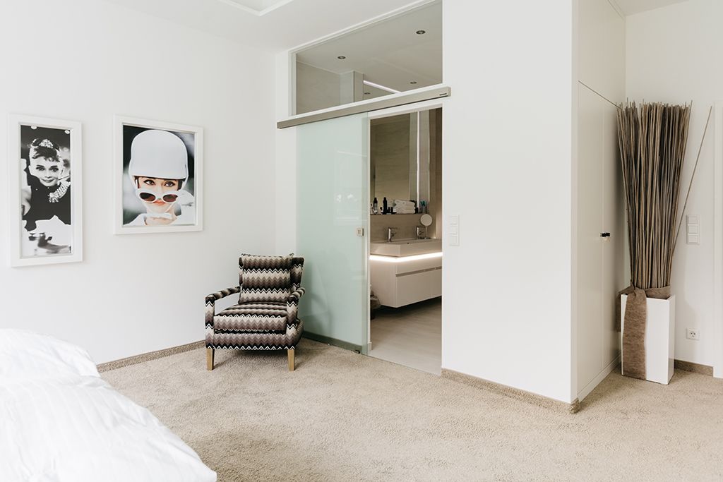 A bedroom with a bed, a patterned armchair, a mirror, and a bathroom visible through an open door.
