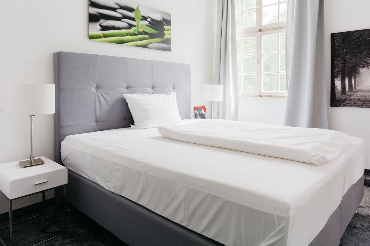 Moderni hotellihuone, jossa on harmaalla kankaalla verhoiltu sänky, valkoiset vuodevaatteet, yöpöytä lampun kanssa ja taideteoksia seinillä.