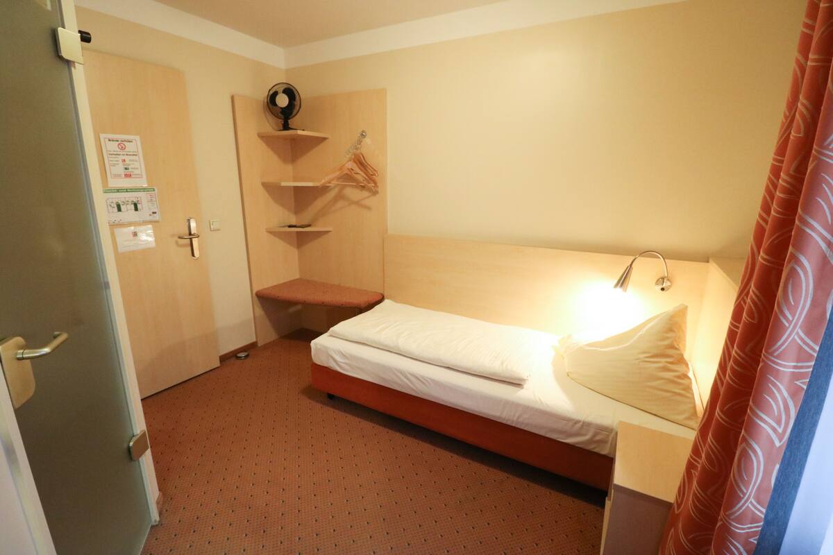 A small hotel room with a single bed, a bedside table, a wall-mounted shelf with a clock, and a door with a handle.