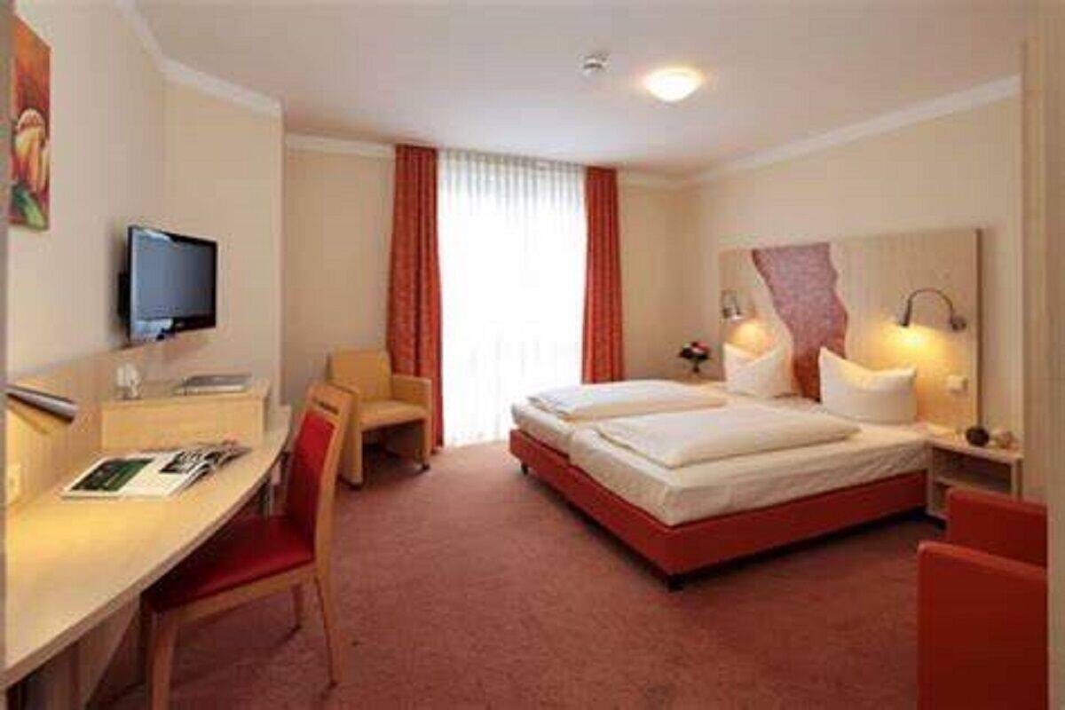 A hotel room with a bed, a desk with a chair, a television, and a window with curtains.