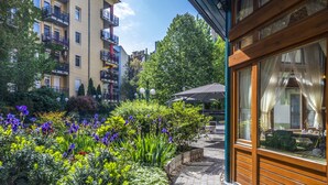 A courtyard with a wooden building, a paved walkway, and a variety of plants and flowers. - Corvin Hotel Budapest - Sissi wing (Budapest)