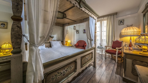 A traditional bedroom with a four-poster bed, a red armchair, and a small table with a lamp. - Corvin Hotel Budapest - Sissi wing (Budapest)