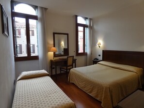 A hotel room with two beds, a desk, a chair, a lamp, a mirror, and two windows with curtains. - Hotel la Forcola (Venice)