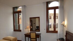 Quarto de hotel com duas camas de solteiro, uma escrivaninha com cadeira, um espelho e uma janela com cortinas.