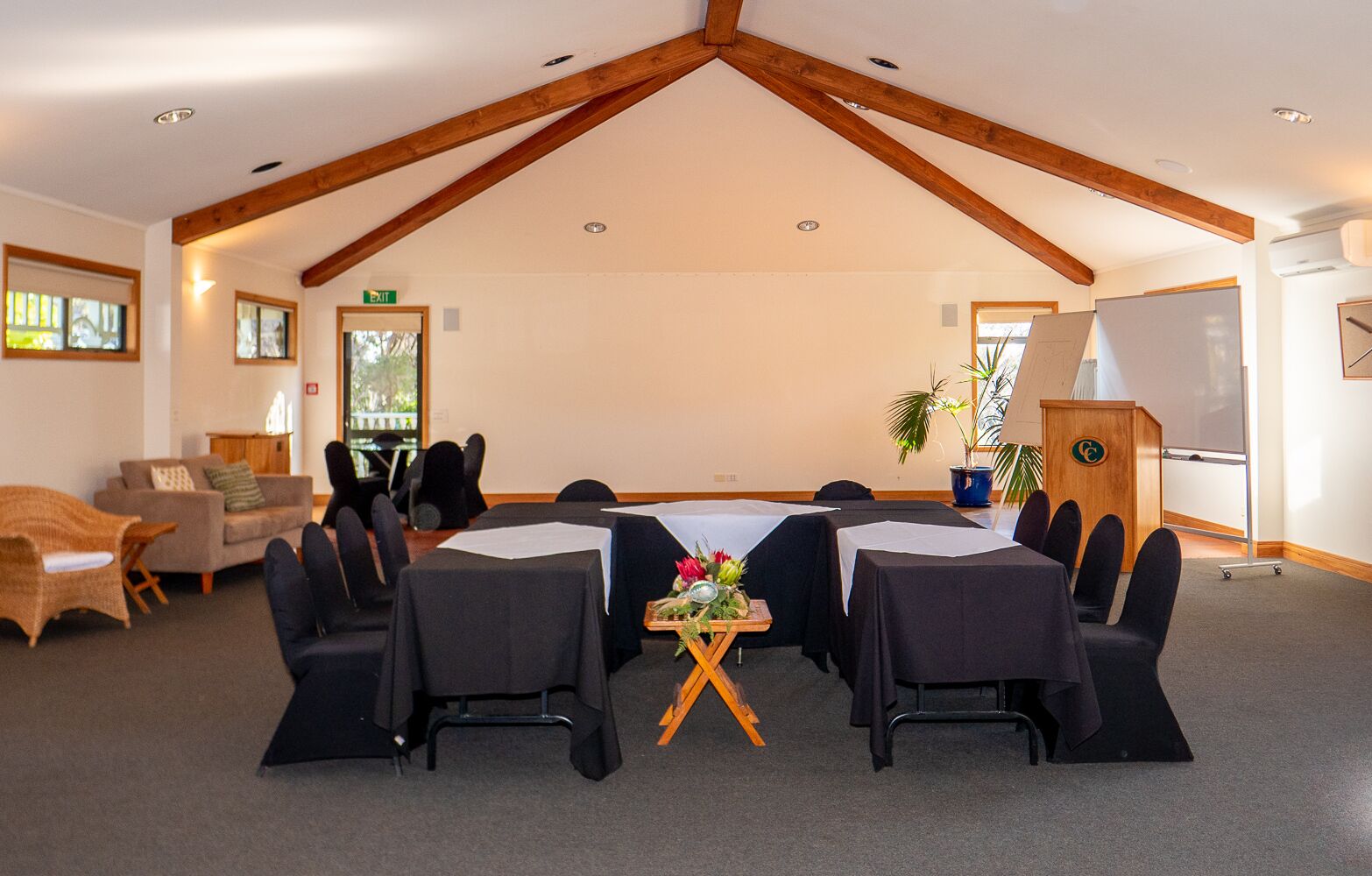 Une salle de conférence dotée d’un plafond haut et de poutres apparentes en bois, aménagée avec des tables et des chaises rondes, un pupitre et un tableau blanc.