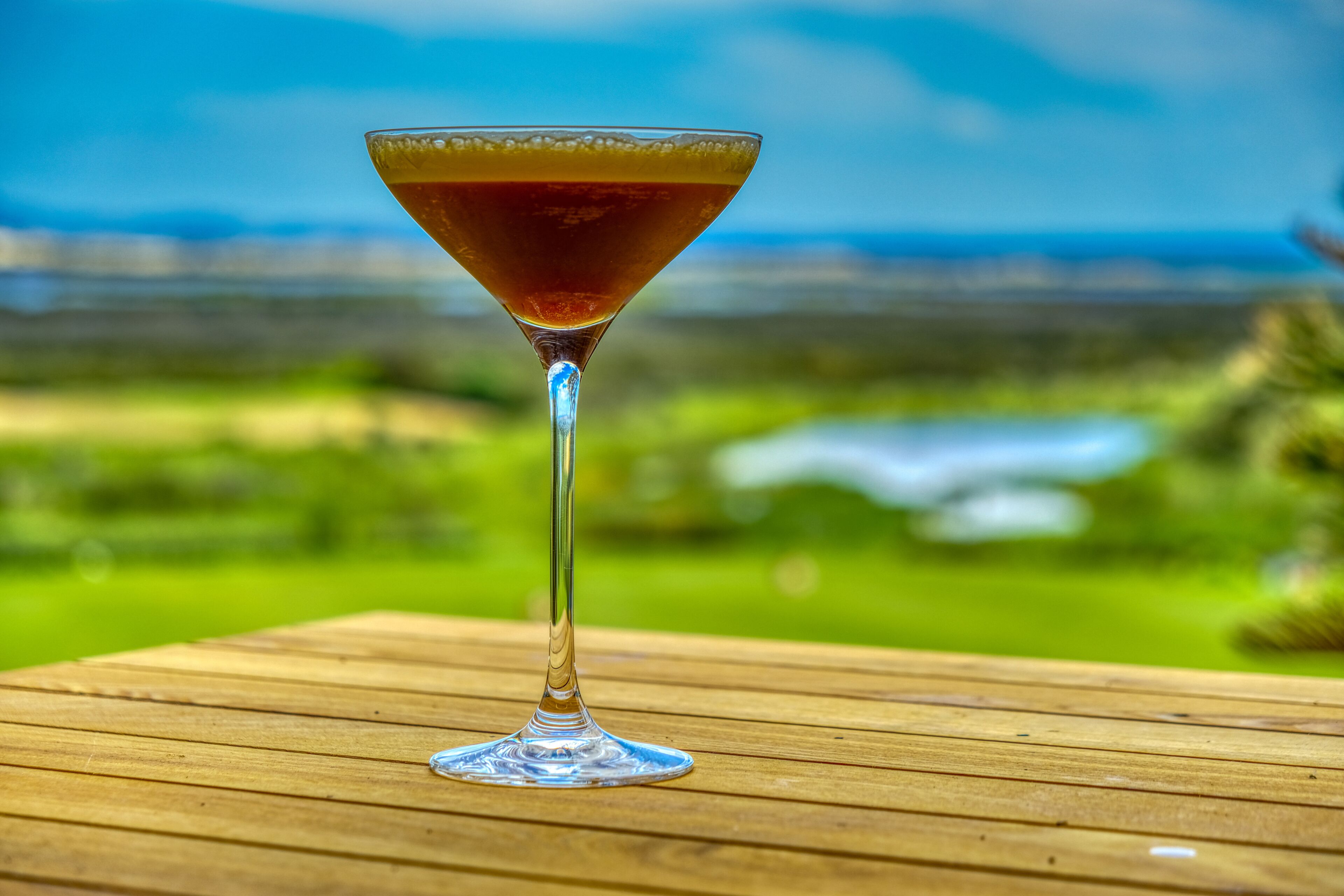 A martini glass on a wooden table with a blurred landscape in the background.