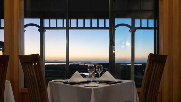 Une table à manger dressée pour deux personnes, avec vue sur le paysage grâce à de grandes fenêtres.