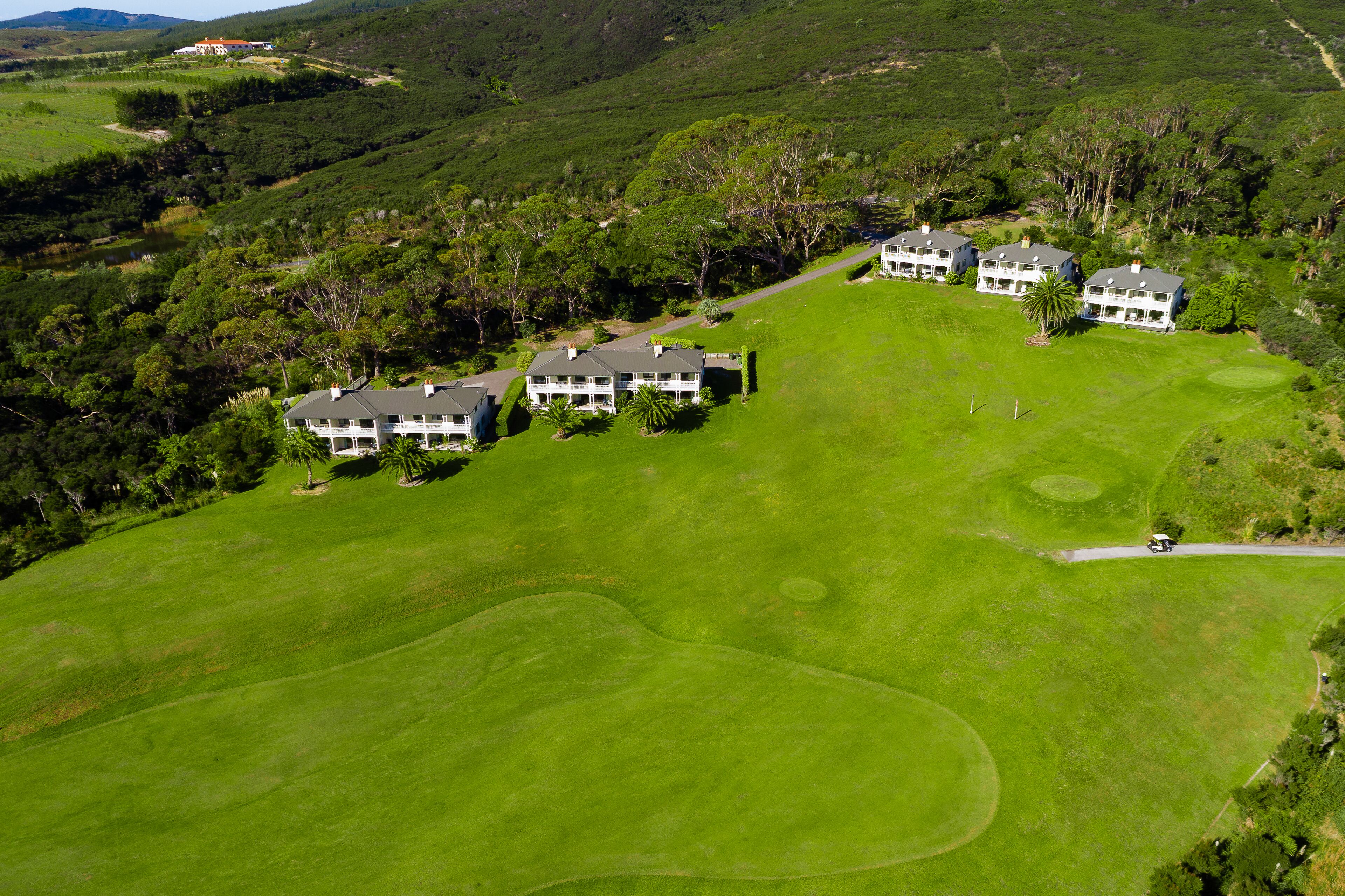 A golf course with multiple buildings, likely a resort or hotel, surrounded by lush greenery and hills.