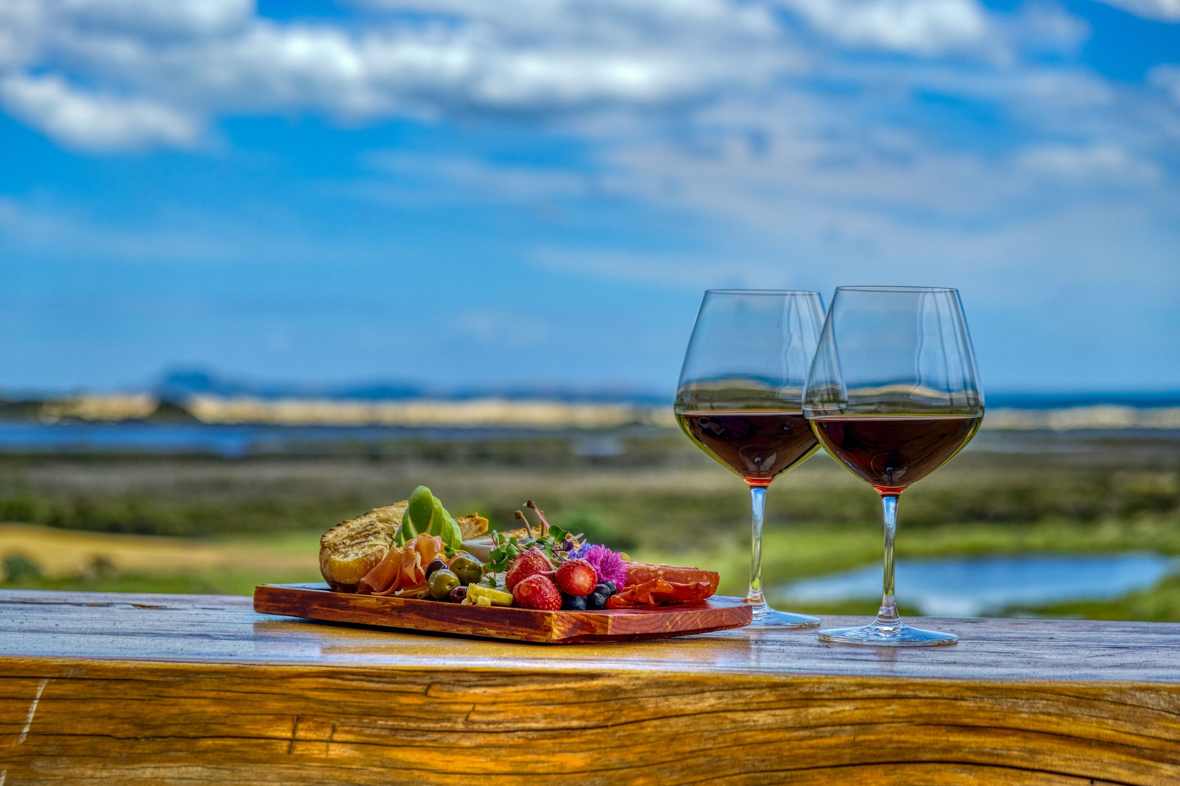 Una mesa de madera con una tabla de embutidos y dos copas de vino frente a un paisaje pintoresco.
