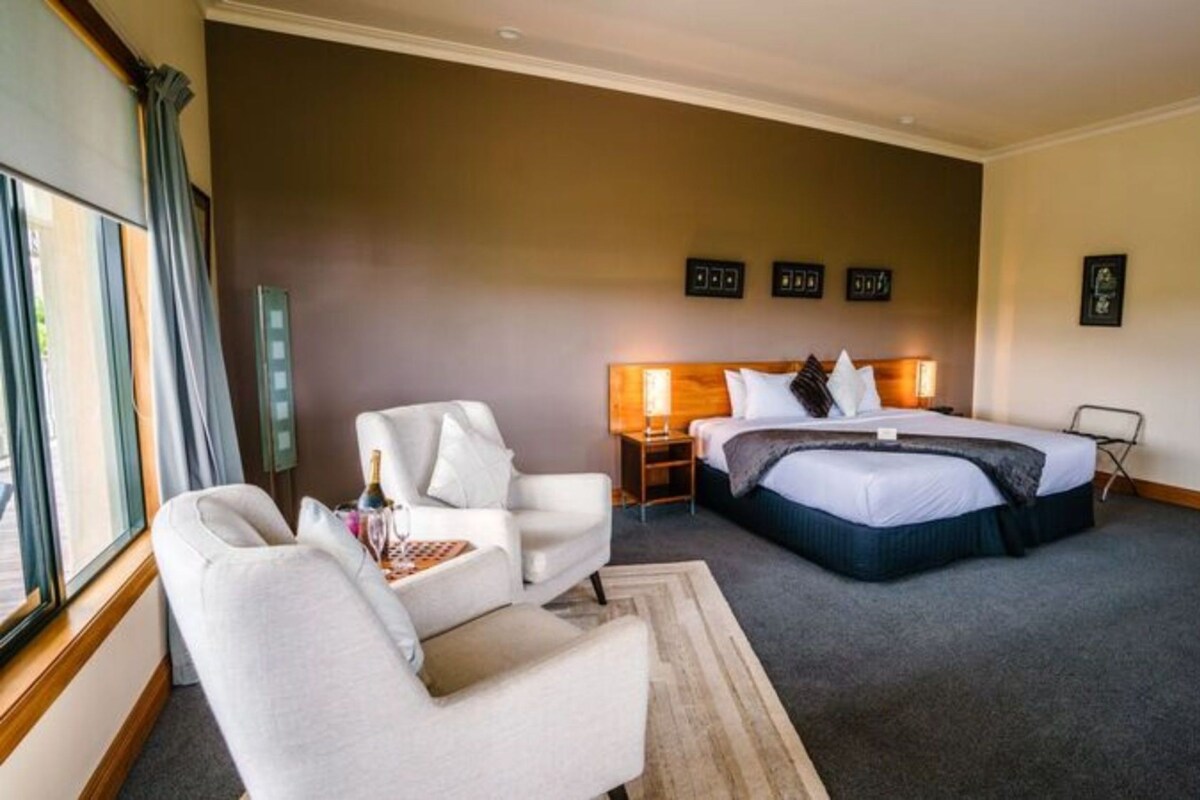 Una habitación de hotel con una cama, dos sillas, una mesita de noche, una lámpara y un ventanal con cortinas.