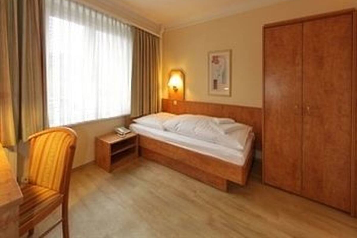 Une chambre d’hôtel comprenant un lit, une table de chevet, une chaise, une armoire et une fenêtre avec des rideaux.