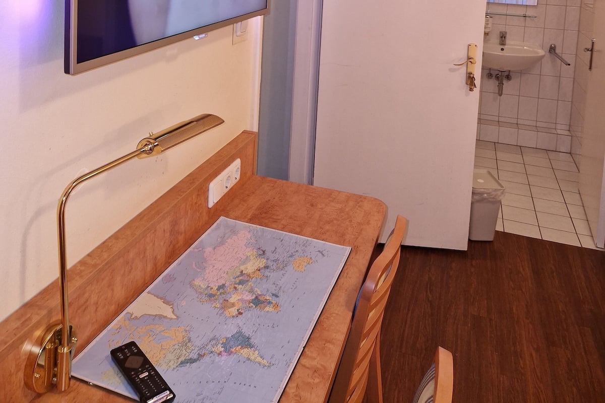 Une chambre d’hôtel dotée d’un téléviseur à écran plat fixé au mur, d’un bureau en bois avec une carte et une télécommande, et d’une salle de bain visible à travers une porte ouverte.