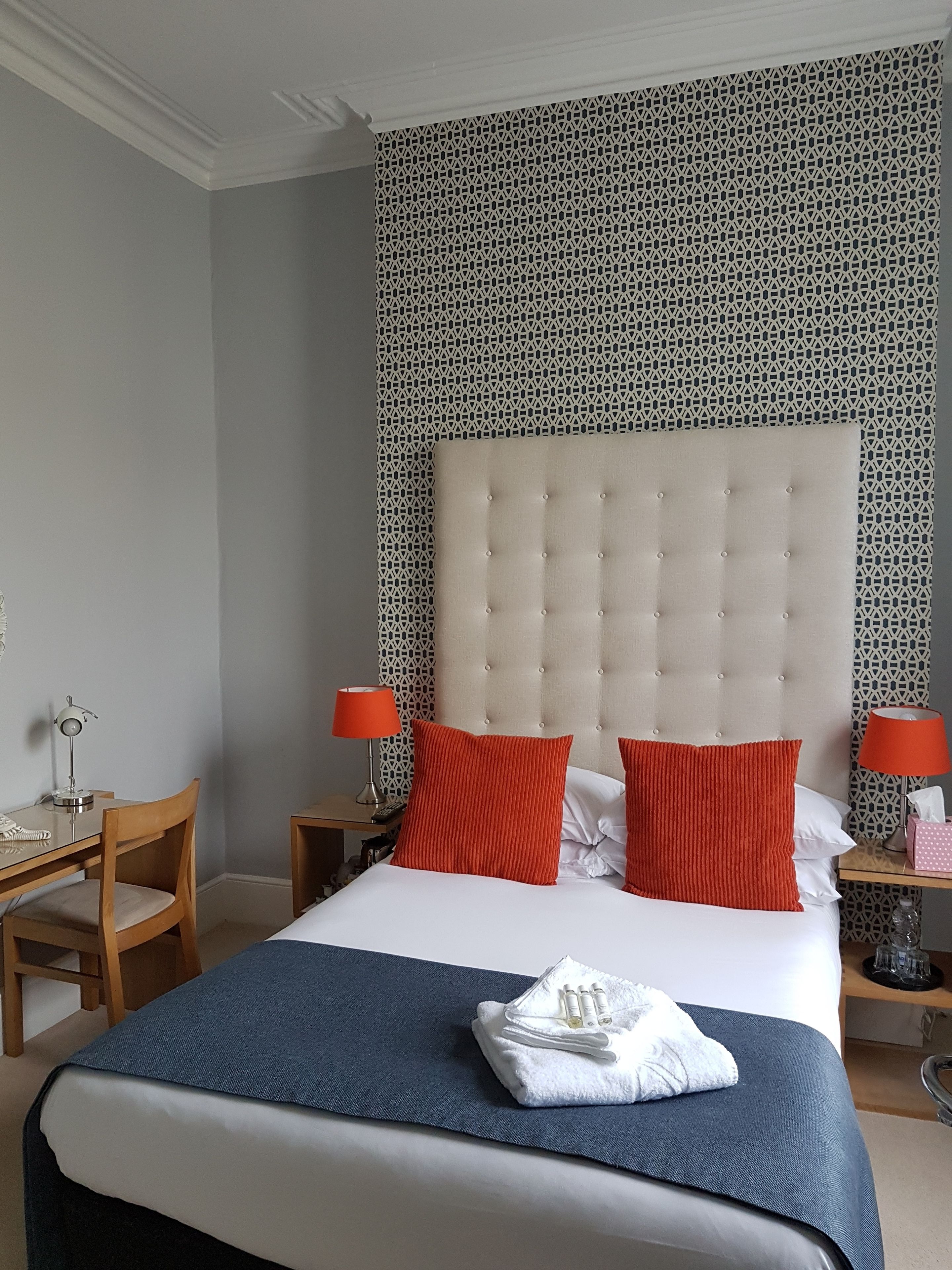 Habitación de hotel con una cama grande, almohadas rojas y blancas, una colcha azul y un papel tapiz estampado.