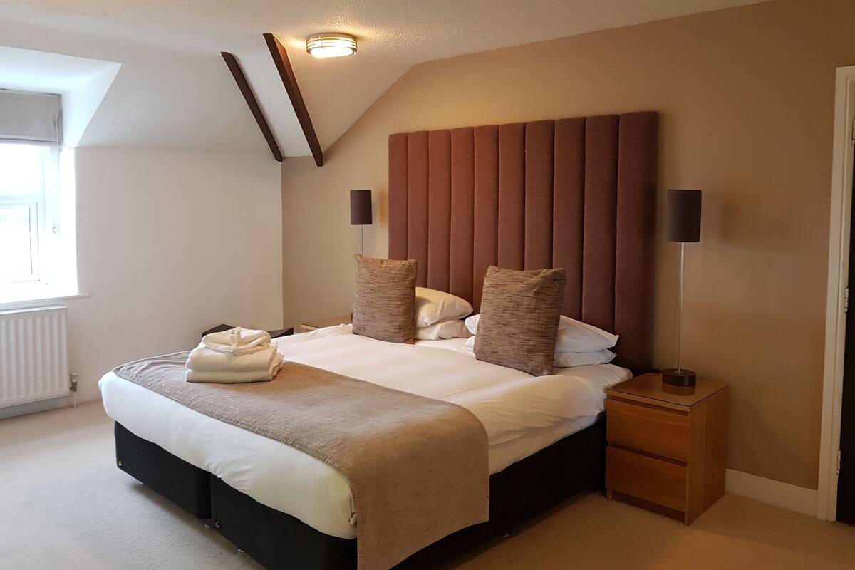 Quarto com cama grande, mesa de cabeceira, duas luminárias na parede e janela com persianas.