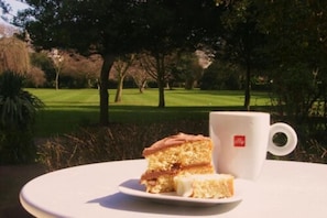 A slice of cake on a plate with a white cup on a round table outdoors.