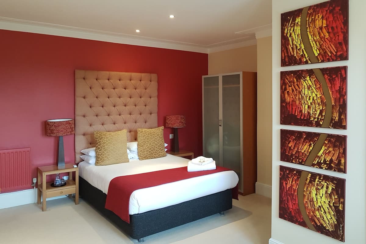 Quarto com cabeceira estofada, cama com roupa de cama vermelha e branca, duas mesas de cabeceira com abajures, um guarda-roupa e um grande quadro abstrato na parede.