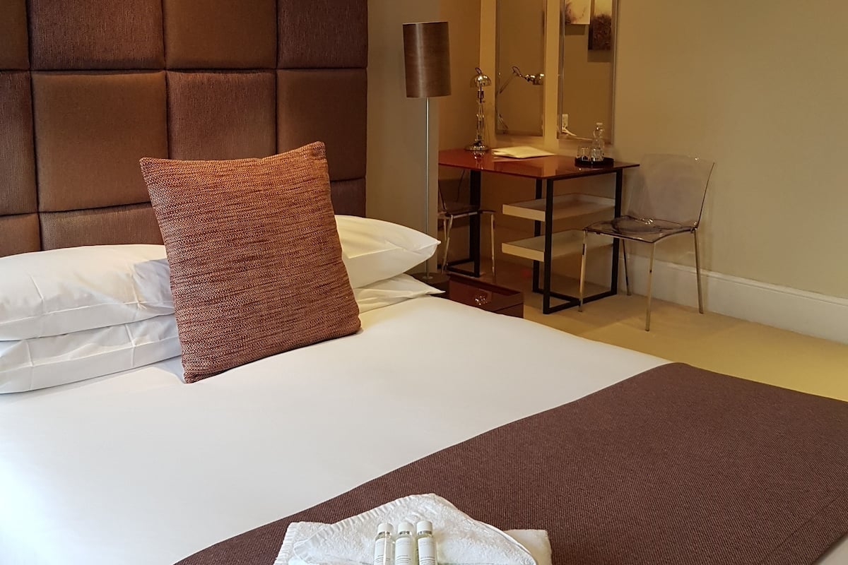 Quarto de hotel com uma cama grande, cabeceira marrom, escrivaninha com espelho e uma cadeira.
