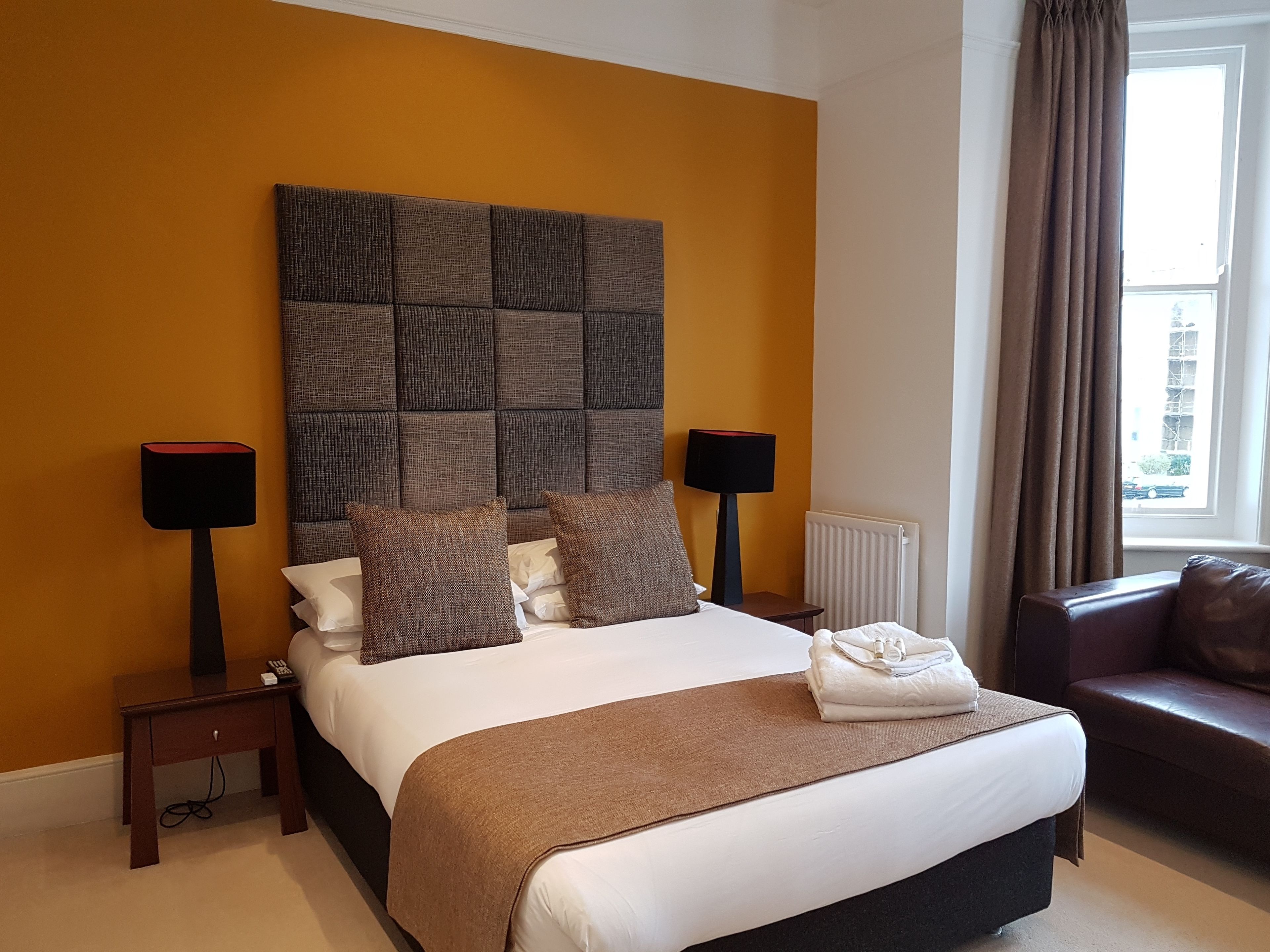 A hotel room with a large bed, two bedside tables, a brown headboard, a window with curtains, and a brown leather sofa.