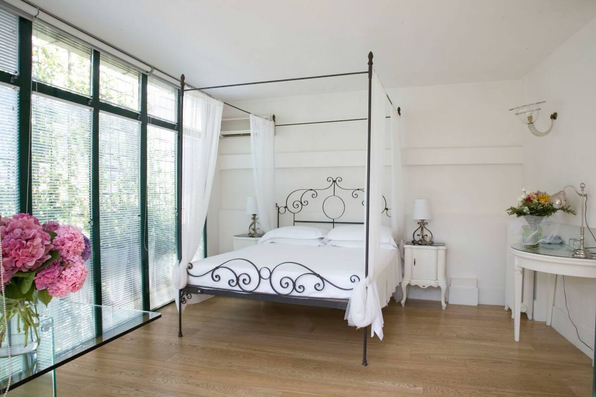 Een slaapkamer met een hemelbed, een nachtkastje met een lamp, een klein wit kastje en een groot raam met gordijnen.