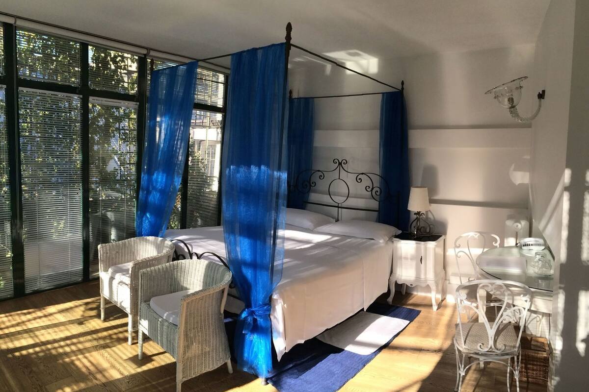 Een slaapkamer met een hemelbed, rieten stoelen, een klein tafeltje en uitzicht door grote ramen.