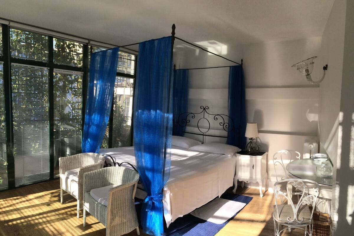 Een slaapkamer met een hemelbed, een rieten stoel, een bijzettafeltje en een glazen pot.