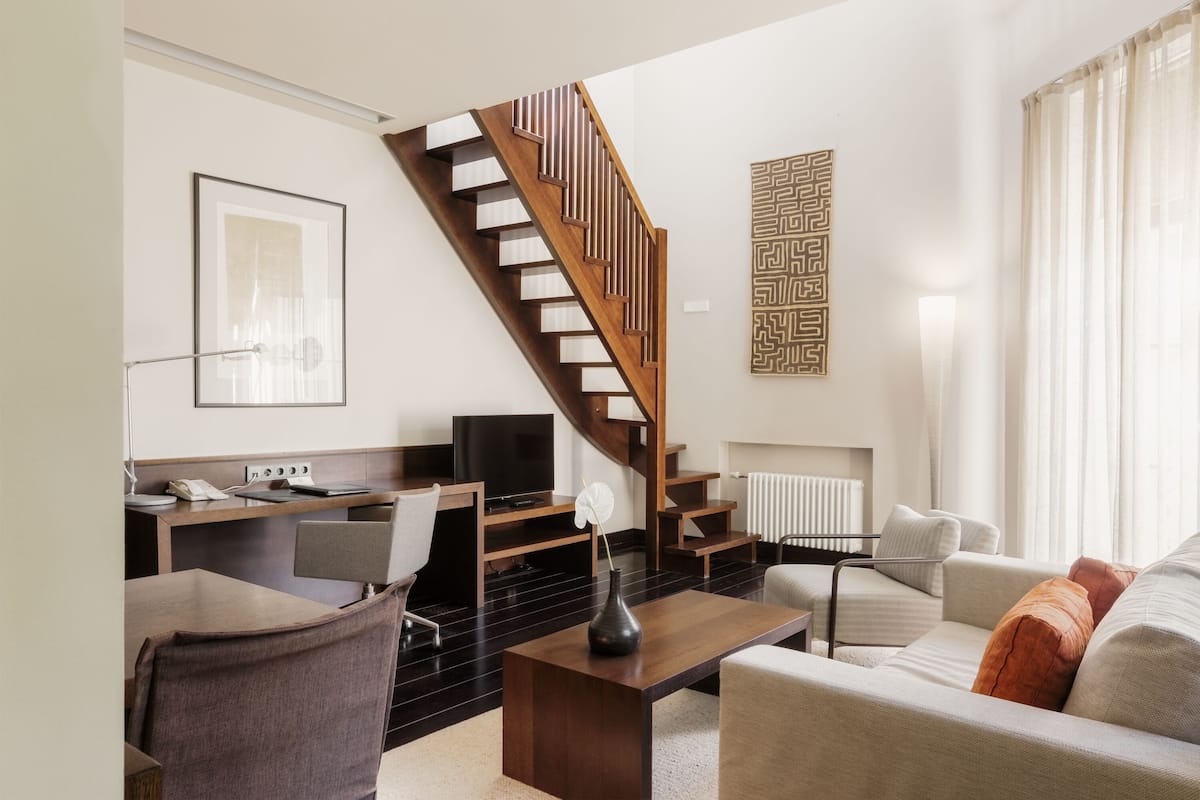 Una sala moderna con una escalera de madera, un televisor de pantalla plana, un comedor y un sofá con cojines.