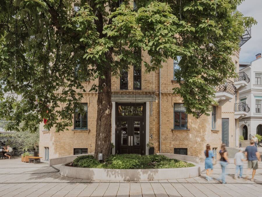 Un edificio con un gran árbol en primer plano, un cartel que dice 'Viesca Hotel Bergs' y una plaza pavimentada con gente caminando.